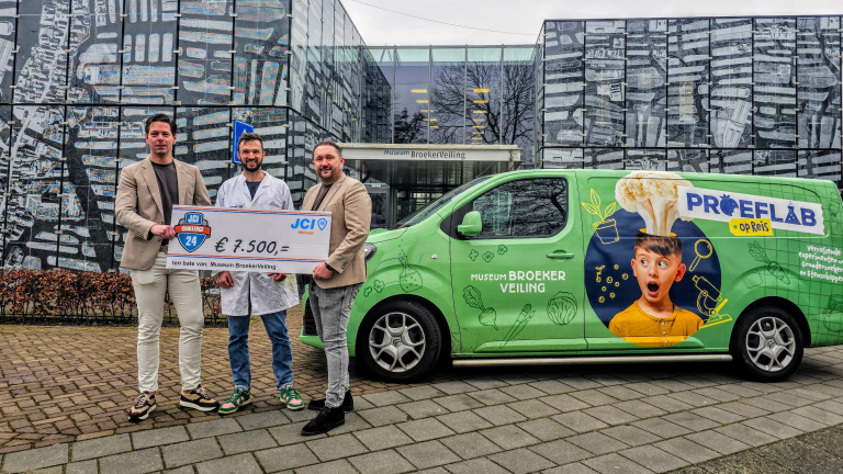 Twee mannen en een wetenschapper houden een cheque vast van €7.500 voor Museum BroekerVeiling, naast een groene bestelwagen van het Proeflab op Reis.