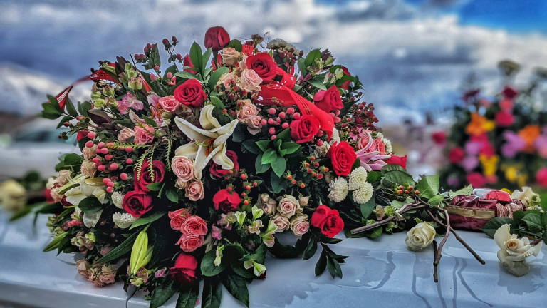 Bloemstuk met rode en roze rozen, lelies en groen blad op een glimmend oppervlak, tegen een bewolkte achtergrond.