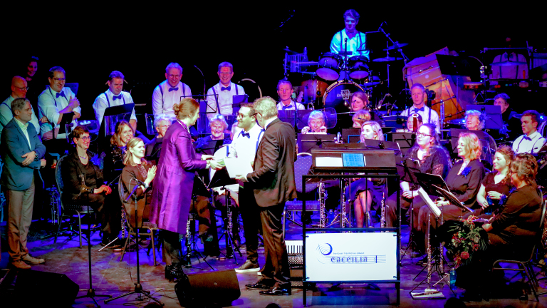 Een orkest op een podium, mensen overhandigen papieren aan elkaar, muzikanten zitten en staan, instrumenten op de achtergrond.