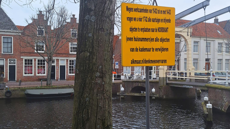 Geel bord met tekst over verplaatsing van voertuigen en objecten wegens werkzaamheden, langs een gracht met historische bakstenen huizen en een brug op de achtergrond.