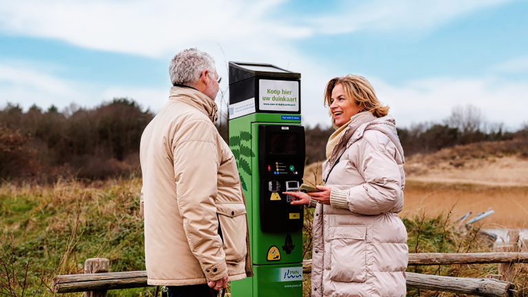 Twee mensen in winterjassen gebruiken samen een groene duinkaart-automaat in een natuurgebied.