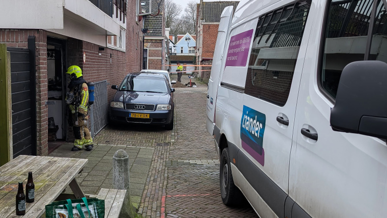 Brandweerman in beschermende kleding bij een deur, geparkeerde auto en Liander-busje op een smalle straat.