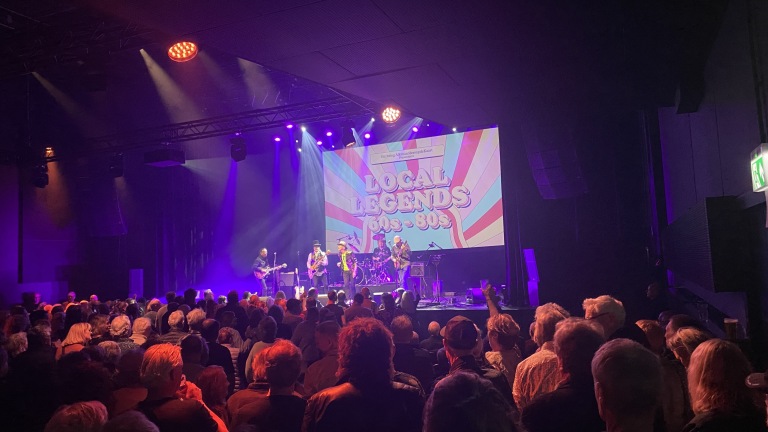 Podiummuziekoptreden met publiek; achtergrond heeft een scherm met tekst "Local Legends 60s-80s."