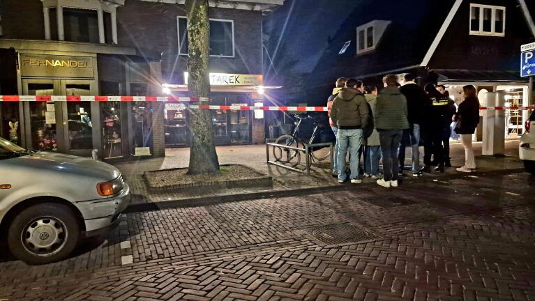 Groep mensen bij een afgezette straat in de avond, met zicht op winkels "Fernandel" en "Tarek".