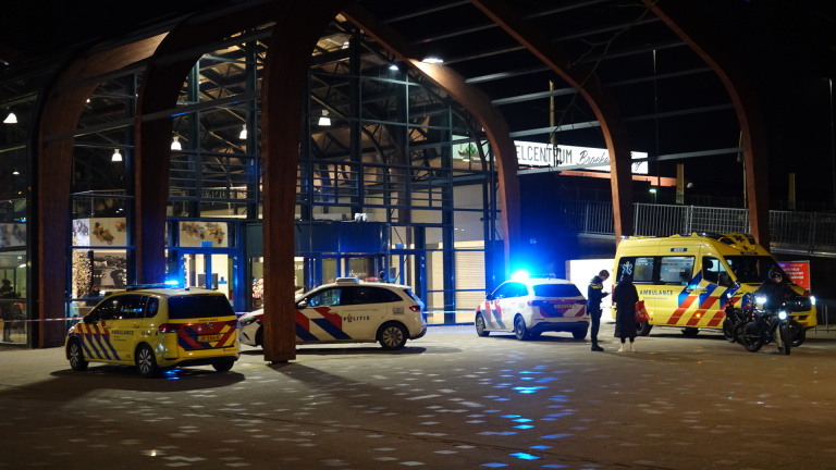 Buitenaanzicht van een gebouw bij nacht met politievoertuigen en een ambulance, blauwe lichten aan, en enkele mensen op de voorgrond.