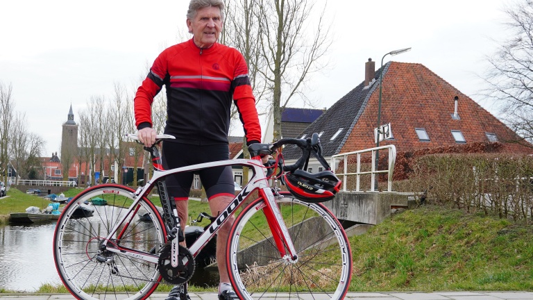 Een man in fietskleding poseert met zijn racefiets naast een kanaal in een dorp met traditionele Nederlandse architectuur.