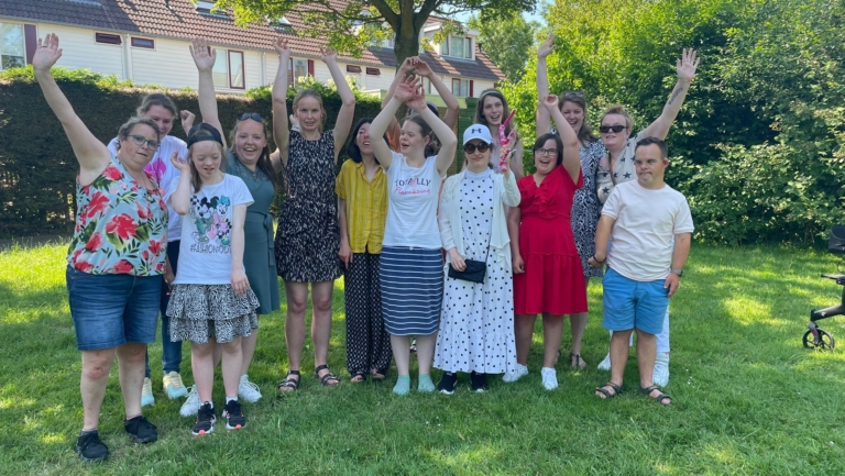 Groep mensen poseert buiten op een grasveld, sommigen met hun handen in de lucht.