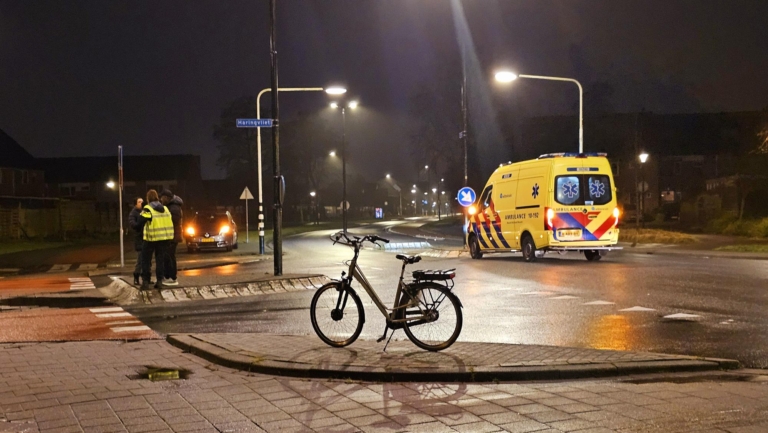 Nachtelijke scène met een ambulance en een fiets op een natte weg; personen in reflecterende vesten staan aan de zijkant.