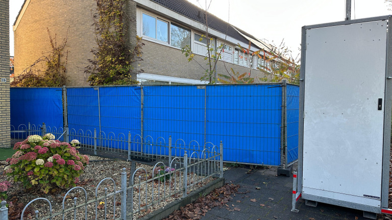 Blauwe bouwhekken naast een huis met een kleine tuin met hortensia's en een mobiel toilet op de stoep.