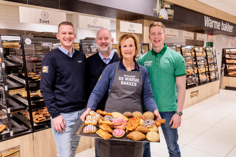 Een groep van vier supermarktmedewerkers poseert in een bakkerijafdeling met een dienblad vol diverse broodjes en gebak.