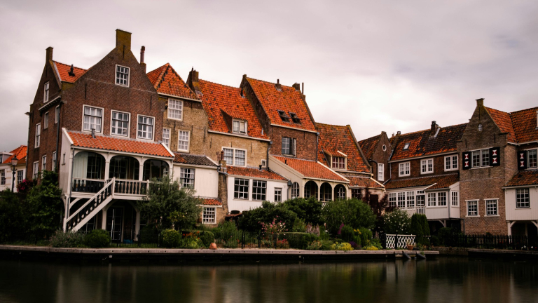 Historische huizen met oranje pannendaken langs een rustige waterkant onder een bewolkte hemel.