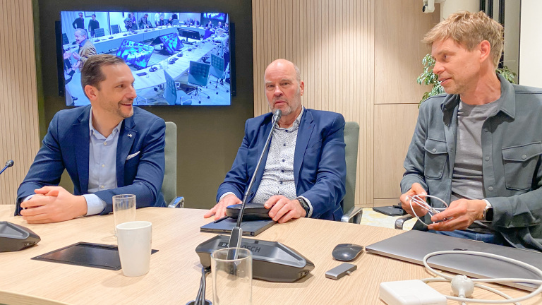 Drie mannen in bespreking aan een vergadertafel met een televisiescherm op de achtergrond dat een andere vergadering toont.