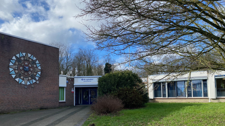 Schoolgebouw met een kunstwerk van mozaïek aan de muur, omgeven door groen gras en bomen, met een ingang met het bord "de spinaker speciaal onderwijs".