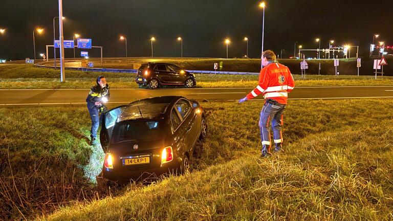 Een auto in de berm met twee hulpverleners erbij op een donkere snelweg, omgeven door straatverlichting.