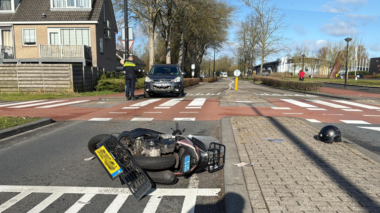 Een omgevallen scooter ligt op de weg bij een kruispunt, met een politieagent en een auto in de achtergrond. Een helm ligt op de stoep naast de scooter.