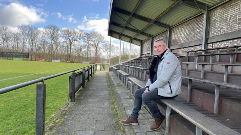 Een man zit op een lege tribune bij een voetbalveld onder een bewolkte blauwe lucht.