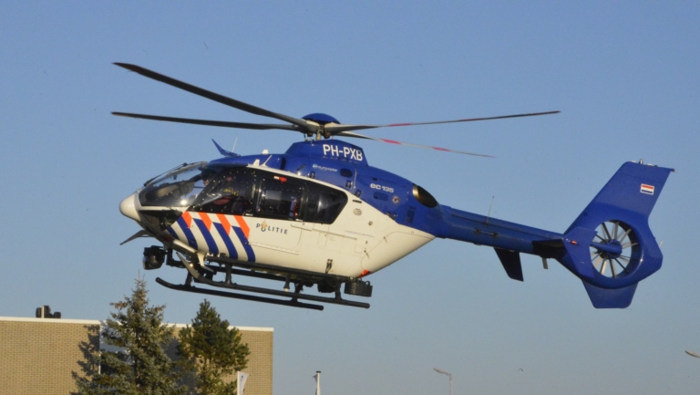 Een Nederlandse politiehelikopter vliegt boven een gebouw, met blauwe lucht op de achtergrond.