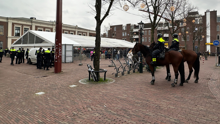 Politieagenten te paard en te voet bij een plein met tenten en wachtrijen.