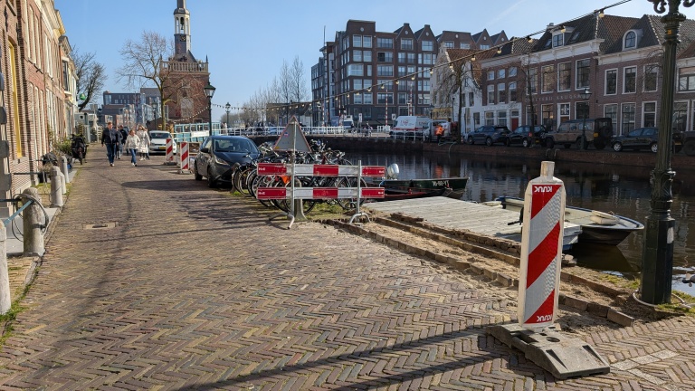 Een straat langs een gracht met geparkeerde fietsen en auto's, afgebakend door rood-witte bouwhekken en borden. Rechts een aanlegsteiger en op de achtergrond een historische toren en moderne gebouwen.