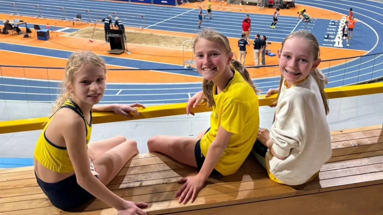 Drie meisjes in sportkleding zitten op houten banken en kijken naar een indoor atletiekbaan.