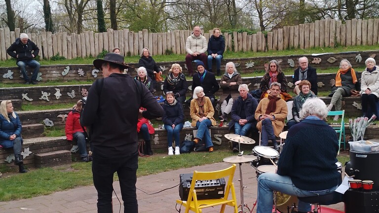 Personen kijken naar een muziekoptreden in een amfitheater met houten tribunes en decoraties.