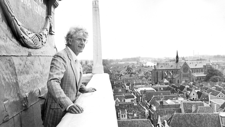 Man in pak kijkt uit over daken van een stad vanaf een hoog uitkijkpunt, met kerk en historisch gebouw in de achtergrond.