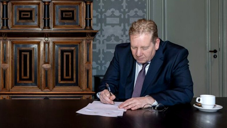 Een man in een pak schrijft aan een tafel met een kop koffie ernaast.