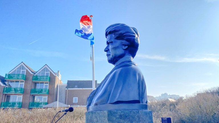 Borstbeeld van een vrouw met de Nederlandse vlag op de achtergrond, naast moderne gebouwen.