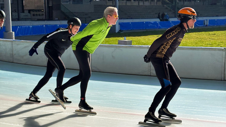 Drie schaatsers op een ijsbaan, met zonlicht dat hun schaduwen werpt.