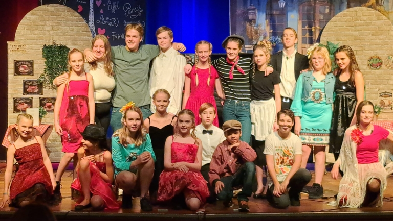 Groep jonge acteurs en actrices op een podium, gekleed in kleurrijke kostuums, poseren samen na een voorstelling. Achtergrond met Franse decoraties.