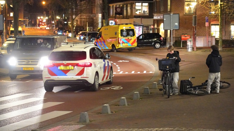 Twee jongeren met fietsen bij een kruispunt met politie- en ambulancevoertuigen.