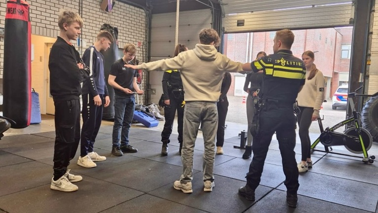 Groep jongeren in een sportschoolomgeving, met agenten van de politie aanwezig. Eén jongere staat met gespreide armen, terwijl anderen toekijken.