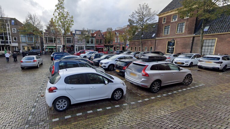 Parkeerplaats op een plein met geparkeerde auto's, omringd door historische gebouwen en enkele bomen, met bewolkte lucht op de achtergrond.