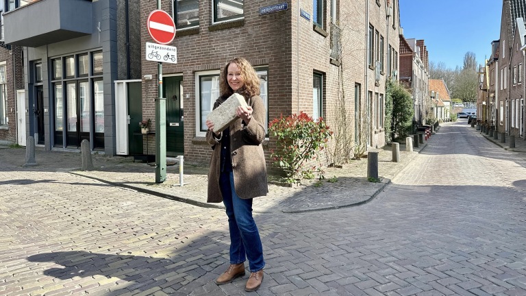 Vrouw met jas houdt een grote steen vast op een geplaveide straat met bakstenen gebouwen en een bord "uitgezonderd fietsers" op de achtergrond.