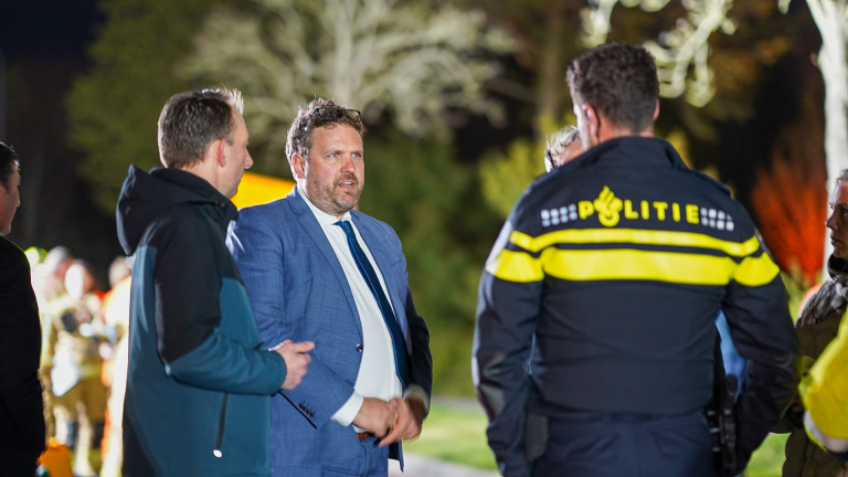 Mensen staan 's avonds buiten in gesprek, waaronder een persoon in een politie-uniform en een man in een blauw pak.