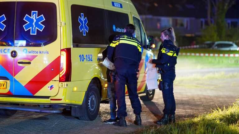 Twee politieagenten staan bij een ambulance met opschrift "witte kruis" terwijl ze papieren vasthouden; het is nacht en er is politieafzetting op de achtergrond.