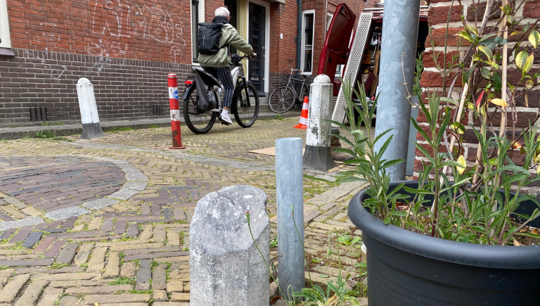 Een persoon op een fiets rijdt langs een rode bestelwagen in een stenen straat met meerdere palen en een zwarte plantenbak met groene planten.