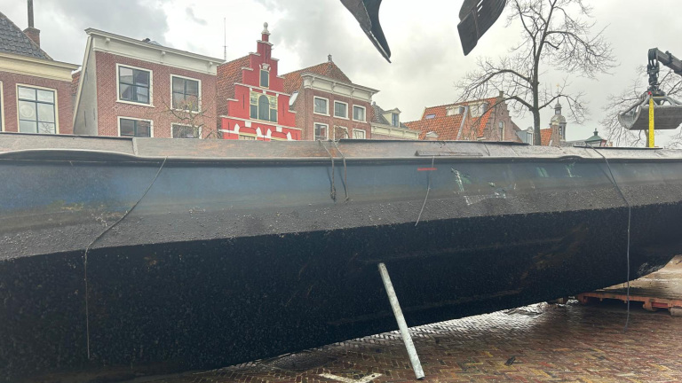 Een omgekeerde, roestige boot ligt op straat tegen de achtergrond van Nederlandse huizen met traditionele bakstenen gevels.
