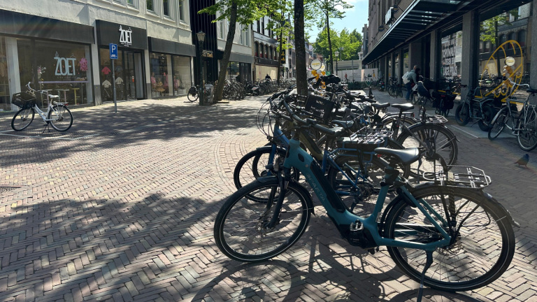 Straatbeeld met geparkeerde fietsen onder bomen, voor een rij winkels met moderne gevels; op de achtergrond is de etalage van een winkel genaamd 'ZOET' zichtbaar.