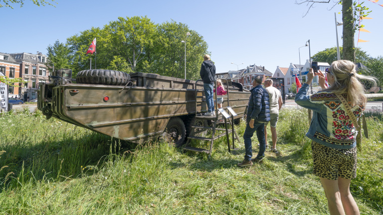 Mensen rond een groot militair amfibievoertuig in een grasveld, met sommige mensen die naar binnen kijken via een ladder en iemand die een foto maakt; stadsgebouwen en bomen in de achtergrond.