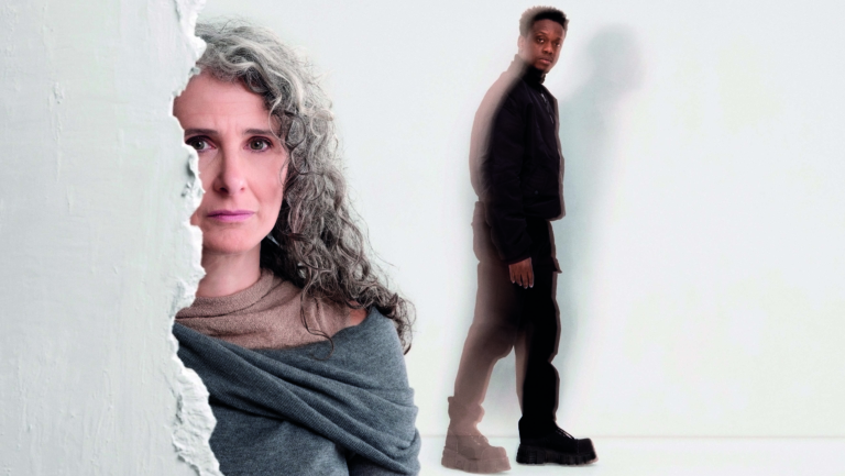 Een vrouw met grijs krullend haar kijkt serieus in de camera, gedeeltelijk verscholen achter een ruwe witte muur, terwijl een man in een donkere outfit verderop loopt met een wazig dubbeleffect.