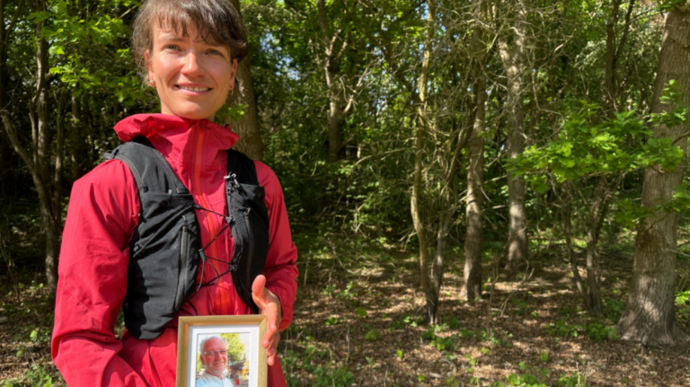 Vrouw in een rood jack staat buiten voor een bos en houdt een ingelijste foto van een oudere man vast.