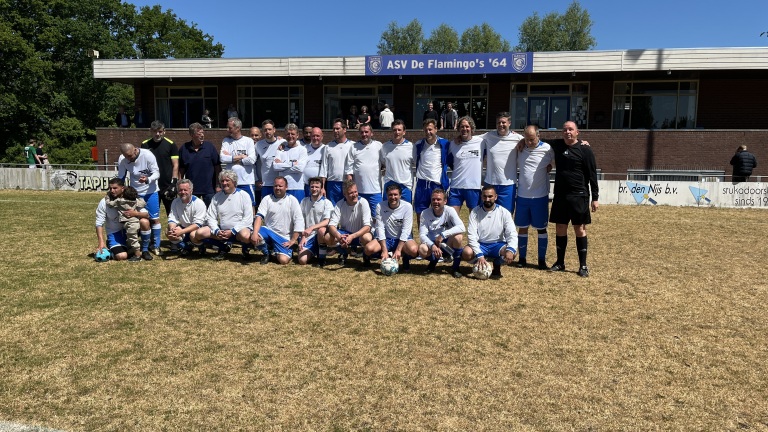 Een groep voetballers poseert voor een teamfoto op een grasveld, gekleed in witte en blauwe tenues, met een bord van ASV De Flamingo's '64 op de achtergrond.
