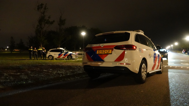 Twee politieauto's met Nederlandse striping staan bij een donkere parkweg. Drie politieagenten zijn te zien in gesprek naast één van de auto's. Het is nacht, en straatlantaarns verlichten de omgeving.