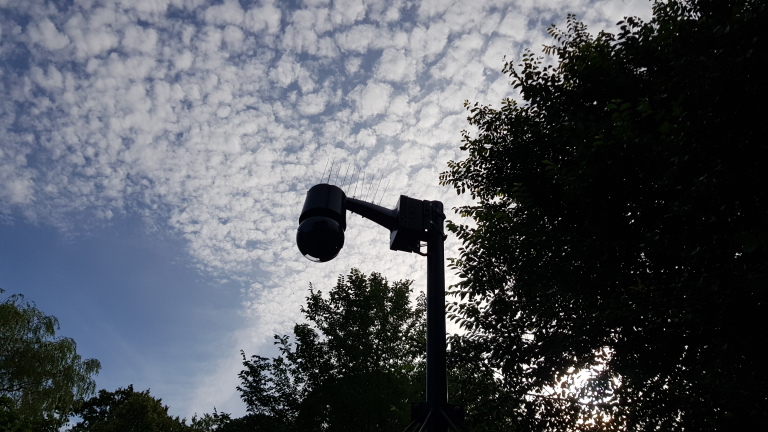 Beveiligingscamera op een paal, omringd door bomen met wolkenlucht op de achtergrond.