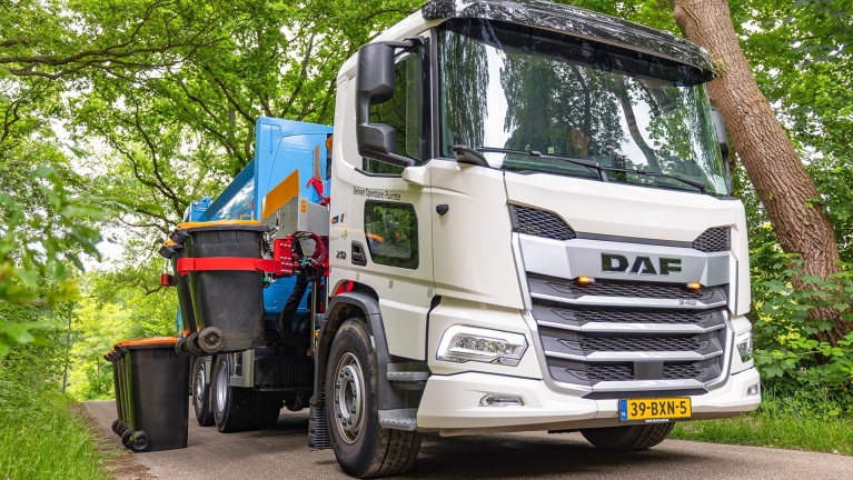 Vuilniswagen van DAF met grijparm die vuilnisbakken optilt, geparkeerd op een smalle weg met bomen aan beide zijden.
