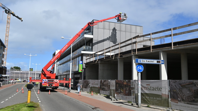 Een rode brandweerwagen met een uitschuifbare ladder voert werkzaamheden uit naast een gebouw in de stad Alkmaar, met een bouwkraan op de achtergrond en een dekkleed met informatie over nieuwe appartementen aan de zijkant van het gebouw.