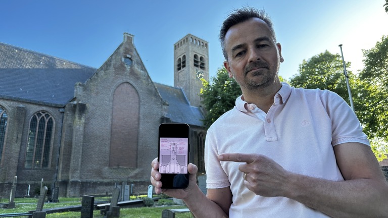Een man in een lichtroze shirt staat buiten voor een historische kerk, terwijl hij naar een mobiele telefoon wijst die hij vasthoudt, waarop een afbeelding van een kerkgebouw te zien is.