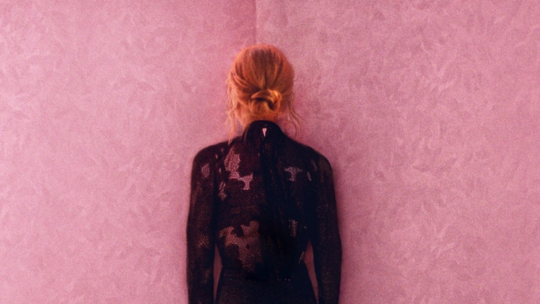 Een persoon met blond haar in een knot staat met de rug naar de camera tegen een roze muur, gekleed in een zwarte jurk met glitterpatroon.