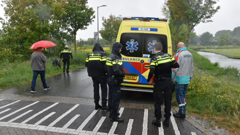 Politieagenten en een ambulance-medewerker staan in de regen naast een ambulance bij een landelijk weggetje; een omstander met rode paraplu loopt weg.